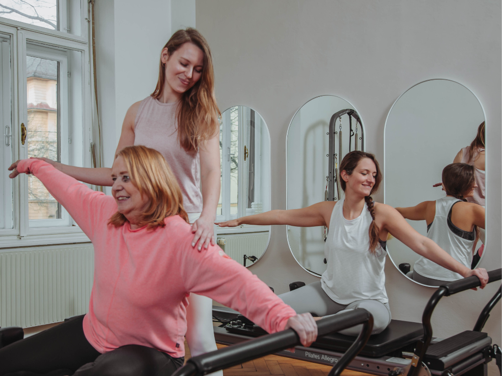 Zwei Frauen während einer Pilates Stunde am Reformer. Die Trainerin unterstützt sie bei den Übungen durch taktile Anleitungen.