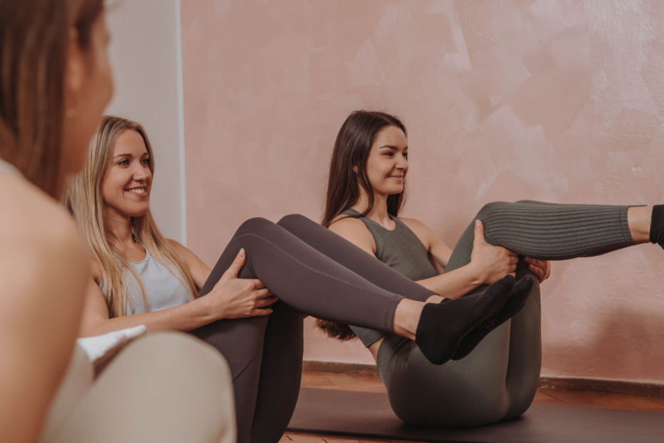 Auf dem Bild sind zwei Frauen in einem Pilates Mattenkurs zu sehen. Sie führen gerade die Pilates Übung Teaser Prep aus. Sie tragen Sportkleidung in Pastell-Farben wie grün, blau und grau.